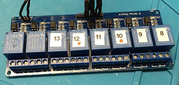 A blue 8-channel relay module with individual blue relays labeled with white stickers numbered 8 to 13, connected by black and red wires at the top, placed on a blue work surface