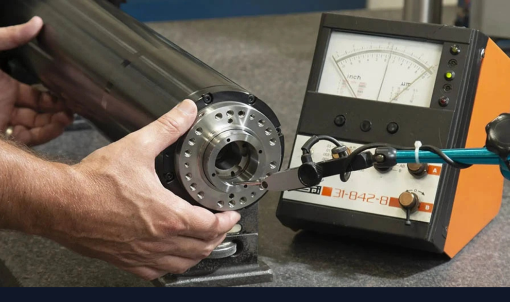 A technician inspects a damaged or worn spindle using a precision dial test indicator, with a measurement device displaying readings in the background. The spindle is mounted on a workbench, and the setup is part of a spindle repair or diagnostic process.